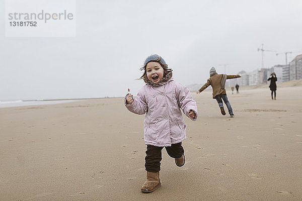 Glückliches kleines Mädchen mit Lolly  das im Winter am Strand läuft  während ihr Bruder im Hintergrund spielt