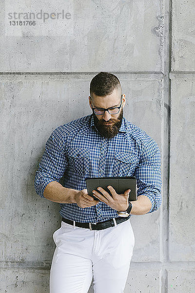 Bärtiger Hipster-Geschäftsmann mit Brille und kariertem Hemd mit Tablette an die Wand gelehnt