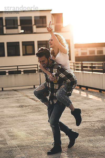 Glücklicher junger Mann  der seine Freundin bei Sonnenuntergang huckepack auf der Dachterrasse mitnimmt