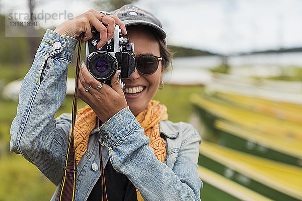 Finnland  Lappland  Porträt einer lächelnden jungen Frau  die mit einer Kamera am Seeufer fotografiert