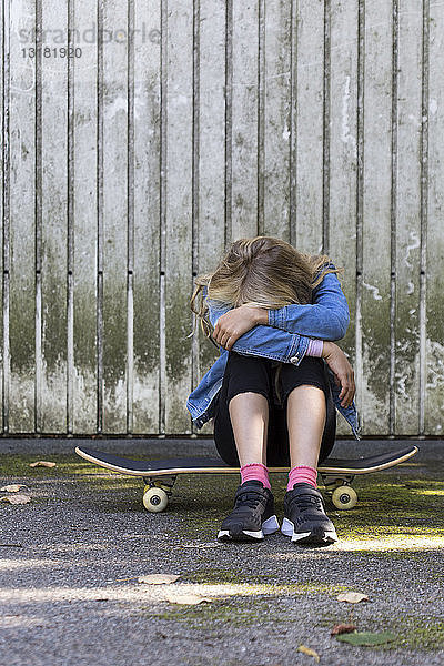 Mädchen sitzt auf ihrem Skateboard im Freien und verbirgt ihr Gesicht