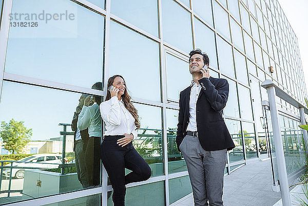 Lächelnde Geschäftsfrau und Geschäftsmann am Handy vor dem Bürogebäude