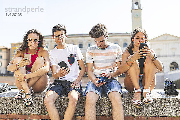 Italien  Pisa  Gruppe von vier Freunden  die gemeinsam mit Mobiltelefonen an einer Wand sitzen