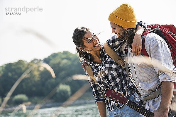 Glückliches junges Paar mit Rucksäcken und Gitarre bei der Umarmung am Flussufer