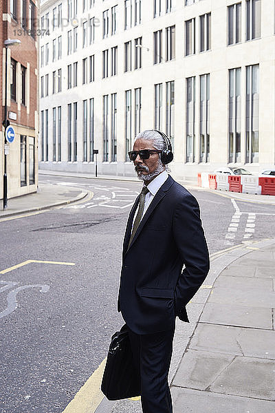 Porträt eines eleganten älteren Geschäftsmannes mit Sonnenbrille und Kopfhörer auf dem Bürgersteig stehend