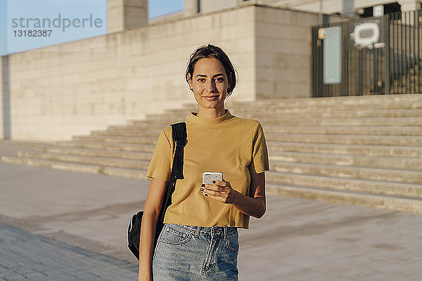 Porträt einer lächelnden jungen Frau  die ihr Handy im Freien in der Hand hält