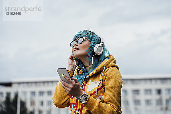 Porträt einer jungen Frau mit blau gefärbten Haaren  die mit Smartphone und Kopfhörern Musik hört