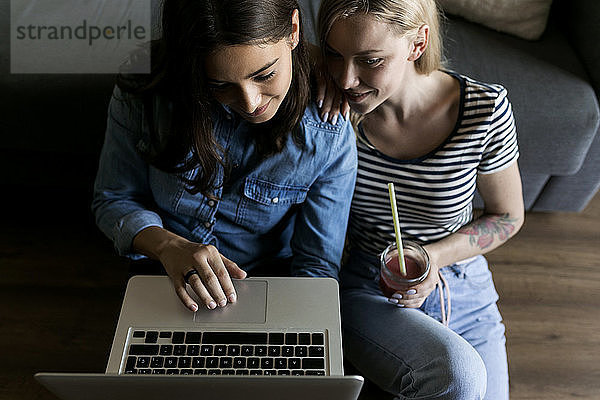 Zwei lächelnde junge Frauen sitzen mit Erfrischungsgetränken auf dem Boden und teilen sich einen Laptop