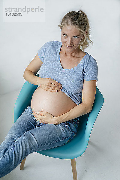 Porträt einer lächelnden schwangeren Frau  die auf einem Stuhl sitzt und ihren Bauch zeigt