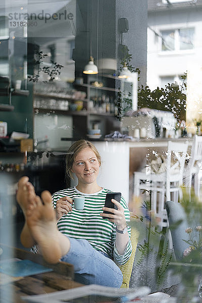 Lächelnde junge Frau mit erhobenen Füßen  die in einem Café Handy und Espressotasse hält