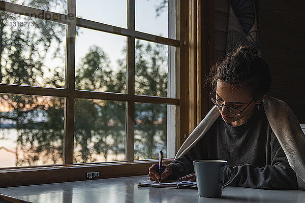 Finnland  Lappland  junge Frau sitzt am Fenster an einem See und schreibt in ihr Tagebuch