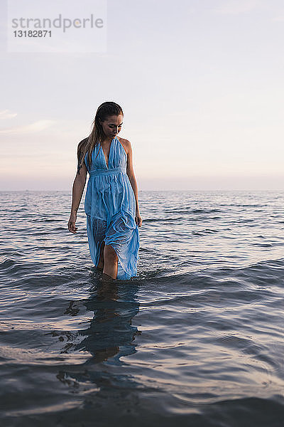 Junge Frau in blauem Kleid geht bei Sonnenuntergang im Wasser am Meeresufer spazieren