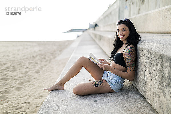 Porträt einer tätowierten jungen Frau  die in der Nähe des Strandes ein Buch liest