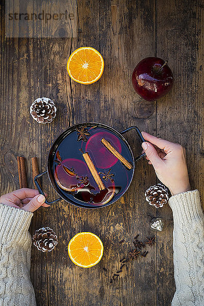 Frauenhände halten einen Glühweinkochtopf mit Orangenscheiben und Gewürzen