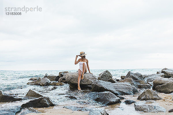 Junge Frau im Badeanzug und mit Hut auf einem Felsen im Meer sitzend