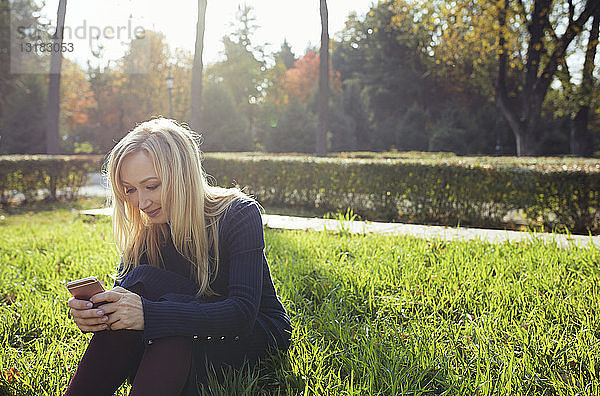 Lächelnde blonde Frau sitzt auf einer Wiese im herbstlichen Stadtpark und benutzt ihr Smartphone