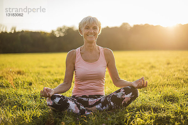 Porträt einer lächelnden älteren Frau in Lotus-Position auf einer ländlichen Wiese bei Sonnenuntergang