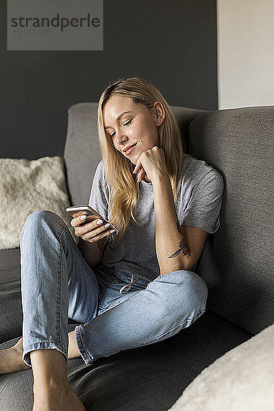 Lächelnde junge Frau sitzt auf Couch und benutzt Mobiltelefon