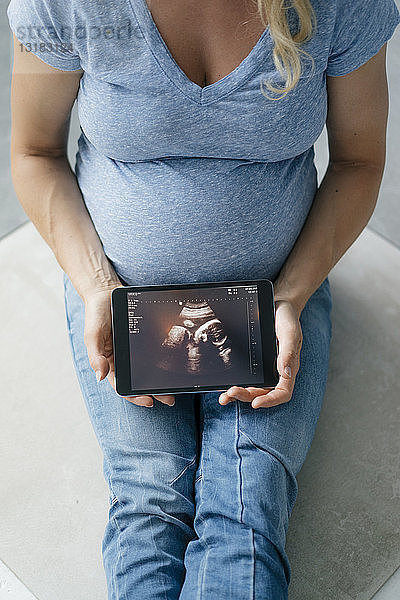 Schwangere Frau sitzt auf dem Boden und zeigt Ultraschallbild auf Tablette