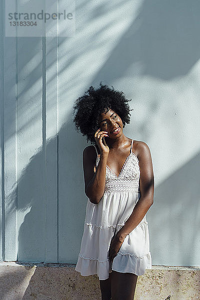 Lächelnde junge Frau in weißem Kleid telefoniert an einer Wand