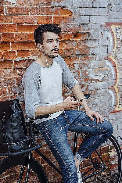 Junger Mann mit Pendler-Fixie-Fahrrad steht mit Handy und Kopfhörern an Ziegelmauer