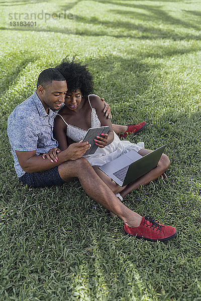 Lächelndes junges Paar teilt sich eine Tablette auf einem Rasen in einem Park