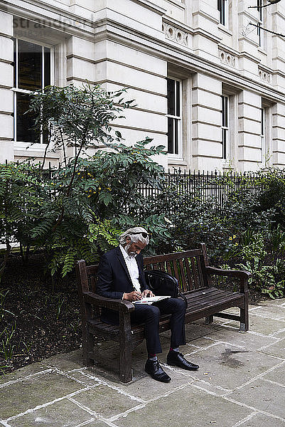 Leitender Geschäftsmann sitzt auf Bank im Freien und schreibt Notizen in Notizbuch