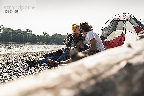 Junges Paar mit Gitarre küsst sich in einem Zelt am Flussufer