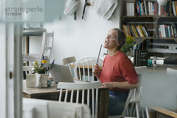 Junge Frau sitzt mit Laptop und Trommelstöcken am Tisch in einem Café