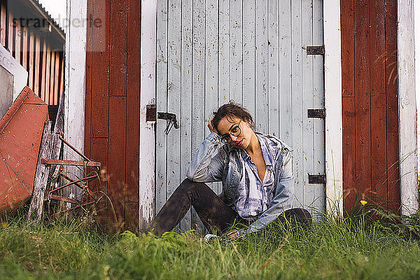 Porträt einer jungen Frau  die im Gras vor einer Holzhütte sitzt