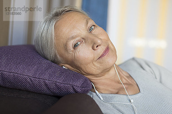 Porträt einer lächelnden älteren Frau  die zu Hause auf der Couch liegt und mit Kopfhörern Musik hört