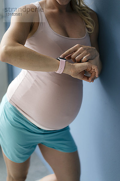 Mittelteil einer schwangeren Frau steht an blauer Wand und schaut auf Smartwatch