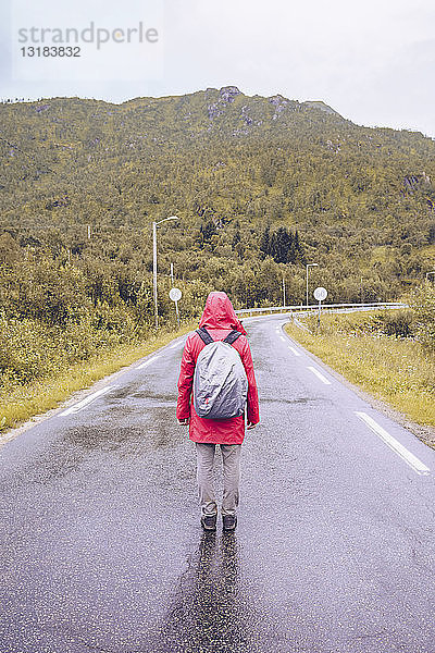 Norwegen  Lofoten  Rückenansicht eines Mannes mit roter Regentasche und Rucksack  der auf nasser Straße steht
