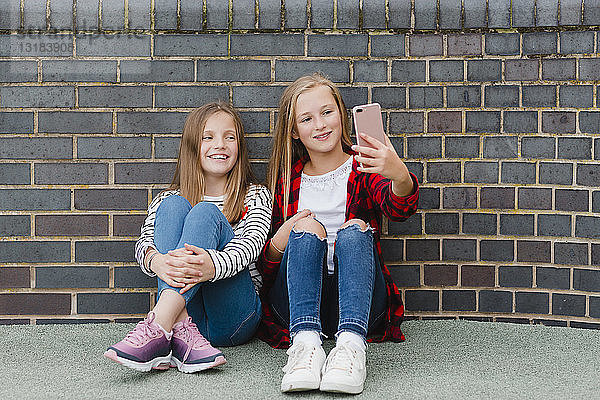 Porträt von zwei lächelnden Mädchen  die vor einer Backsteinmauer sitzen und sich mit einem Smartphone selbstständig machen