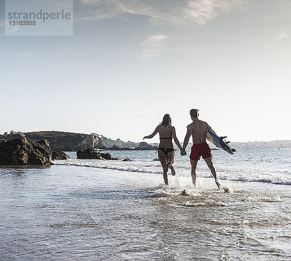 Frankreich  Bretagne  junges Paar mit einem Surfbrett  das Hand in Hand im Meer läuft