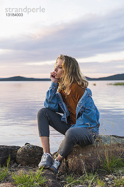 Finnland  Lappland  junge Frau sitzt auf einem Felsen am Seeufer