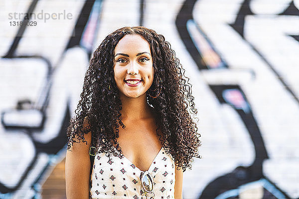 Porträt einer lächelnden jungen Frau mit langen lockigen Haaren an einer Graffiti-Wand