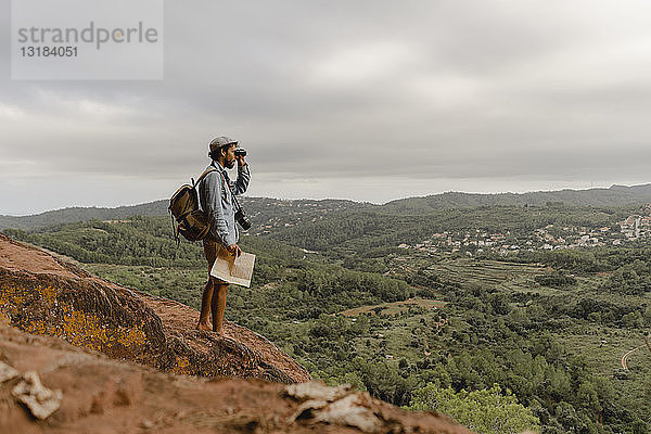 Junger Mann mit Rucksack  auf einem Berg stehend  auf die Aussicht schauend  mit Fernglas