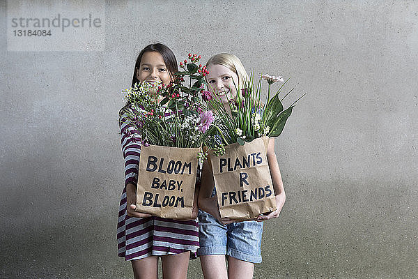 Porträt von zwei lächelnden Mädchen  die nebeneinander stehen und Papiertüten mit Blumen anbieten