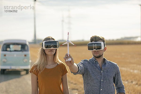 Junges Paar mit VR-Brille am Wohnmobil in ländlicher Landschaft hält Modell einer Windkraftanlage