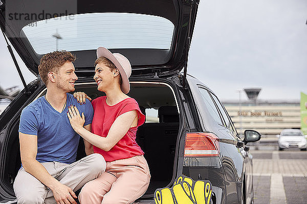 Glückliches junges Paar sitzt im Kofferraum eines Autos auf dem Flughafen