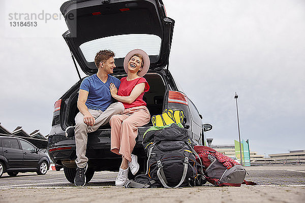 Glückliches junges Paar sitzt im Kofferraum eines Autos auf dem Flughafen
