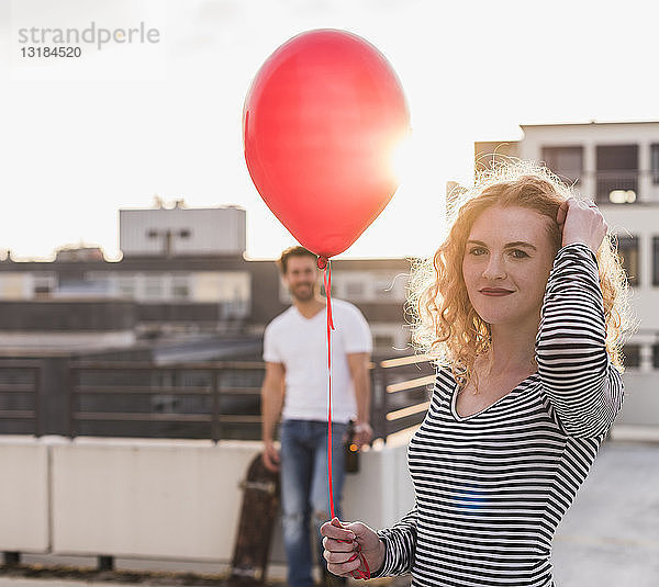 Porträt eines lächelnden jungen Wo-Mannes mit rotem Ballon auf der Dachterrasse bei Sonnenuntergang