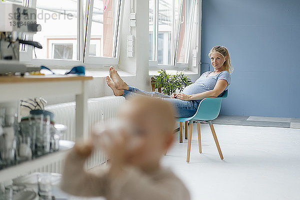 Schwangere Frau sitzt auf einem Stuhl am Fenster mit einem Kleinkind im Vordergrund