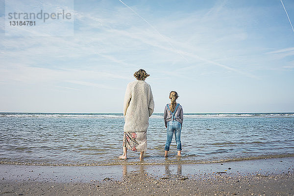 Mutter und Tochter am Strand stehend  auf das Meer blickend  Rückansicht