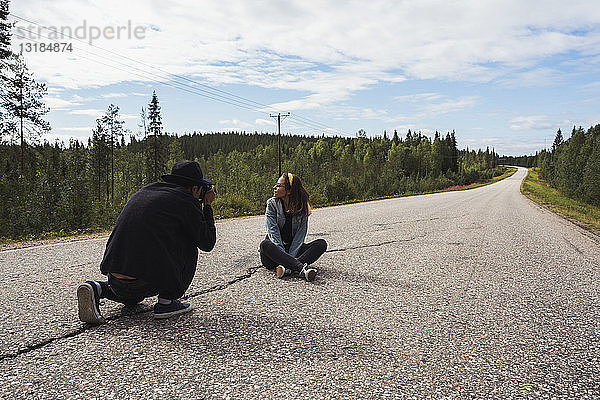 Finnland  Lappland  Mann beim Fotografieren einer Frau  die auf einer leeren Landstraße sitzt