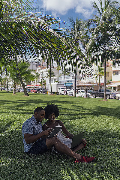 USA  Florida  Miami Beach  junges Paar mit Tablet und Laptop auf einem Rasen in einem Park