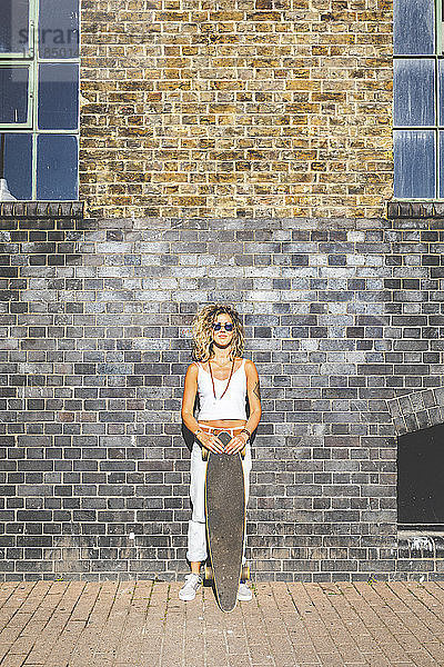 Junge Frau steht mit Skateboard an Ziegelmauer in der Stadt