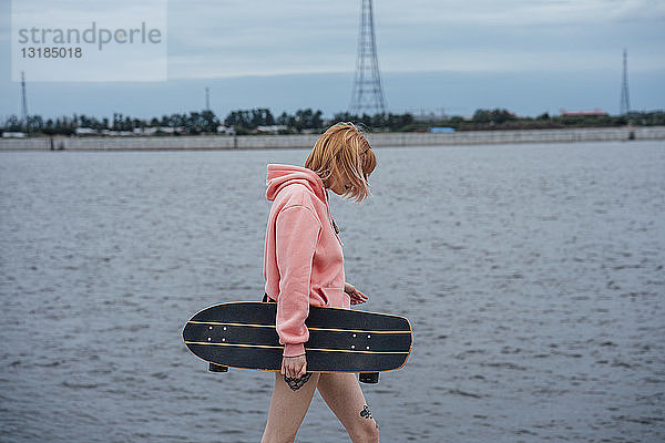 Junge Frau hält Carver Skateboard in der Hand und geht am Flussufer