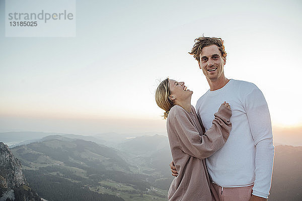 Schweiz  Grosse Mythen  Porträt eines glücklichen jungen Paares  das bei Sonnenaufgang in einer Berglandschaft steht
